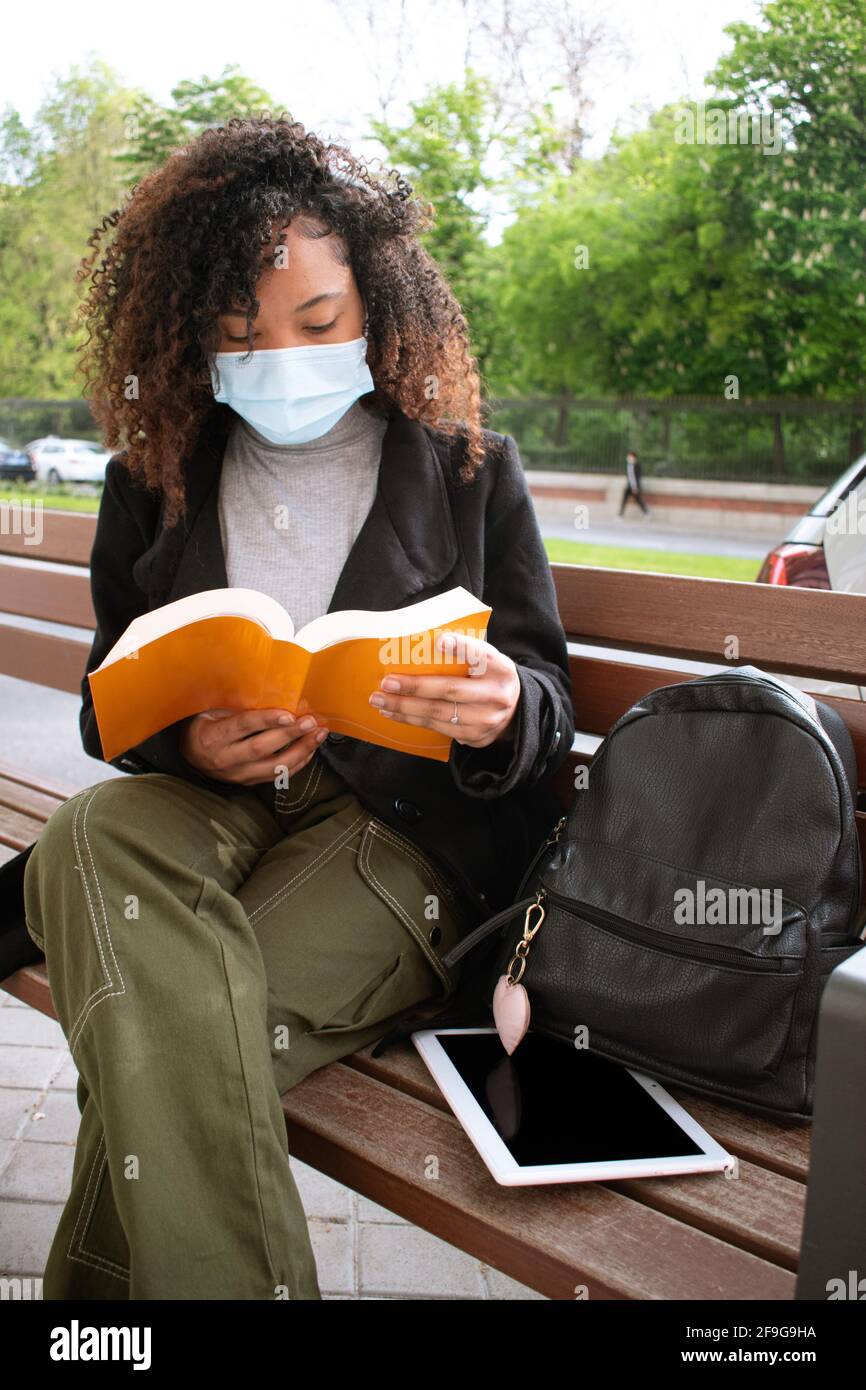 Young woman with curly hair wearing a face mask and reading an orange book on a bench next to her are a backpack and a tablet. Stock Photo