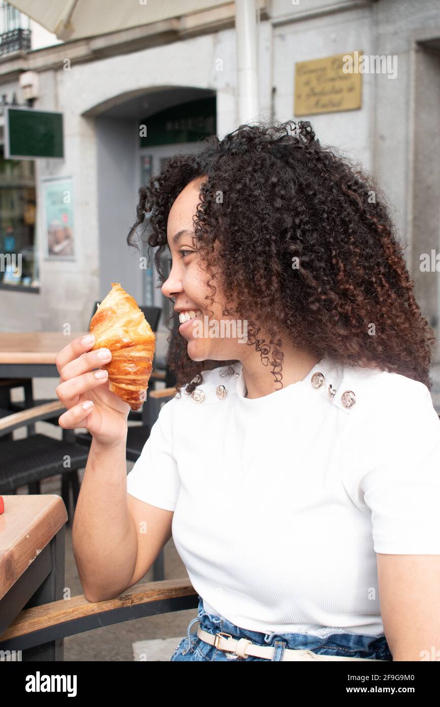 Young woman with curls smiling happily while having a croissant on the terrace of a cafeteria. Stock Photo