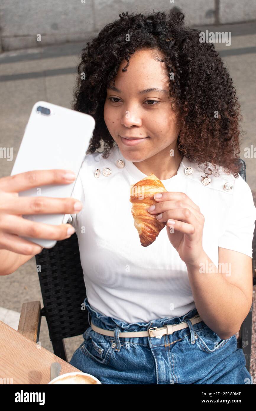 Young black woman with curls eating a croissant while taking a selfie outside a cafe. Stock Photo