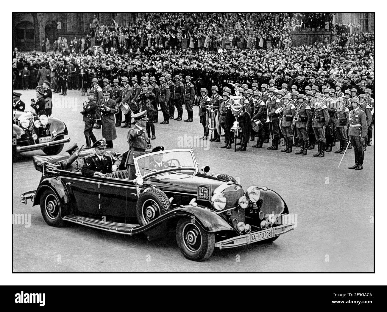 Adolf Hitler in uniform, standing in his official open top Mercedes motorcar wearing swastika armband gives Heil Hitler salute to the military and crowds at a huge Nazi rally in 1938 Germany. Hitler salutes attendees at a Reichsparteitag (Reich Party Day) in Nuremberg, Germany. A uniformed Martin Bormann and Dr Goebbels also in attendance sitting in rear of open top Mercedes Car Stock Photo