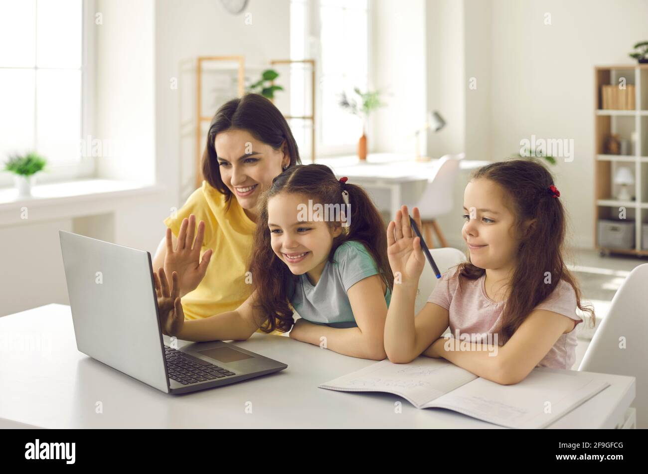 Mom and her two daughters sit in front of a laptop and wave a hand greeting the teacher. Stock Photo