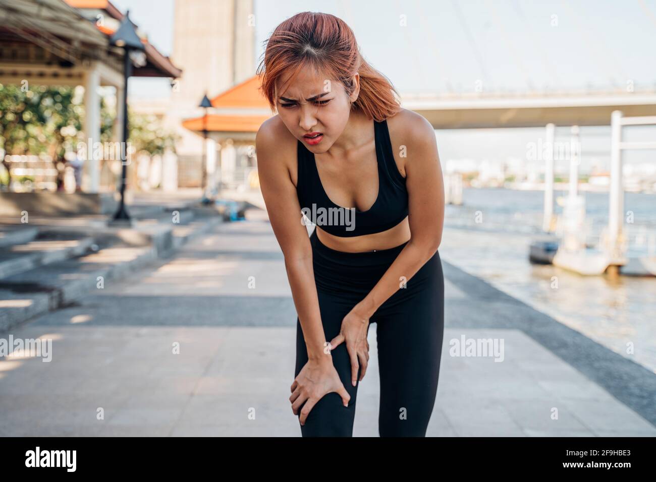 Young Asian female athlete runner in sportswear at river side suffering pain and discomfort from knee injury during morning run Stock Photo