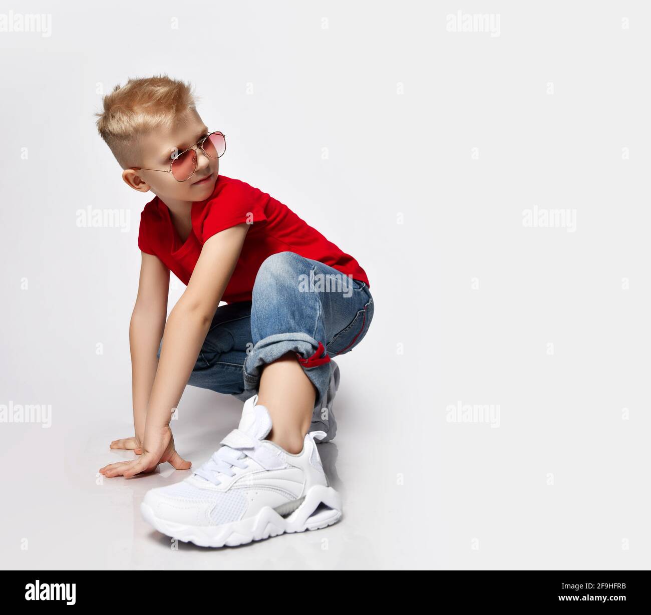 Active, frolic blonde kid boy in red t-shirt, blue jeans, white sneakers and sunglasses is in ready to run position Stock Photo
