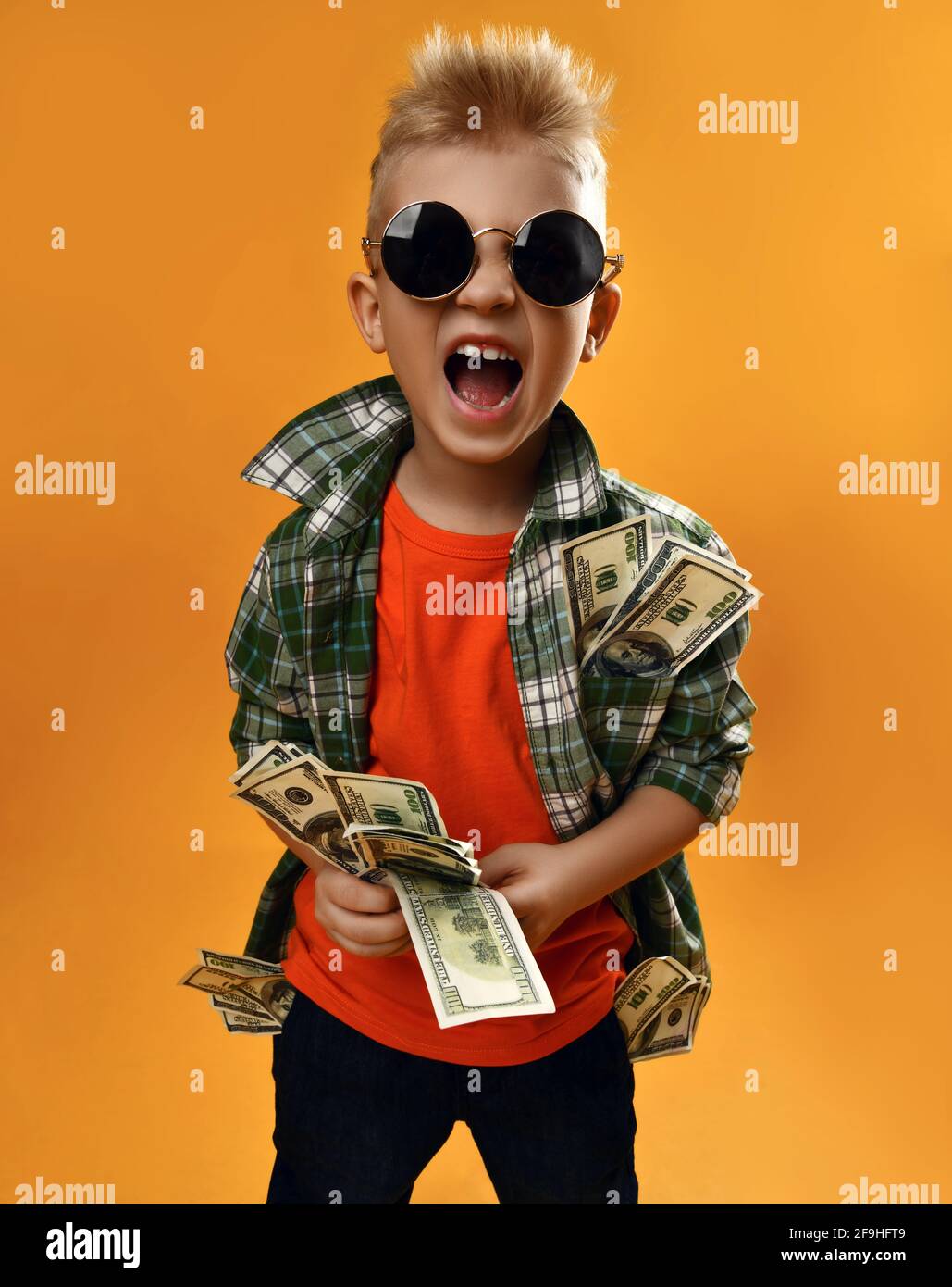 Rich kid boy in round sunglasses, checkered plaid shirt and jeans stands with bundle of dollar cash in hands and screams Stock Photo