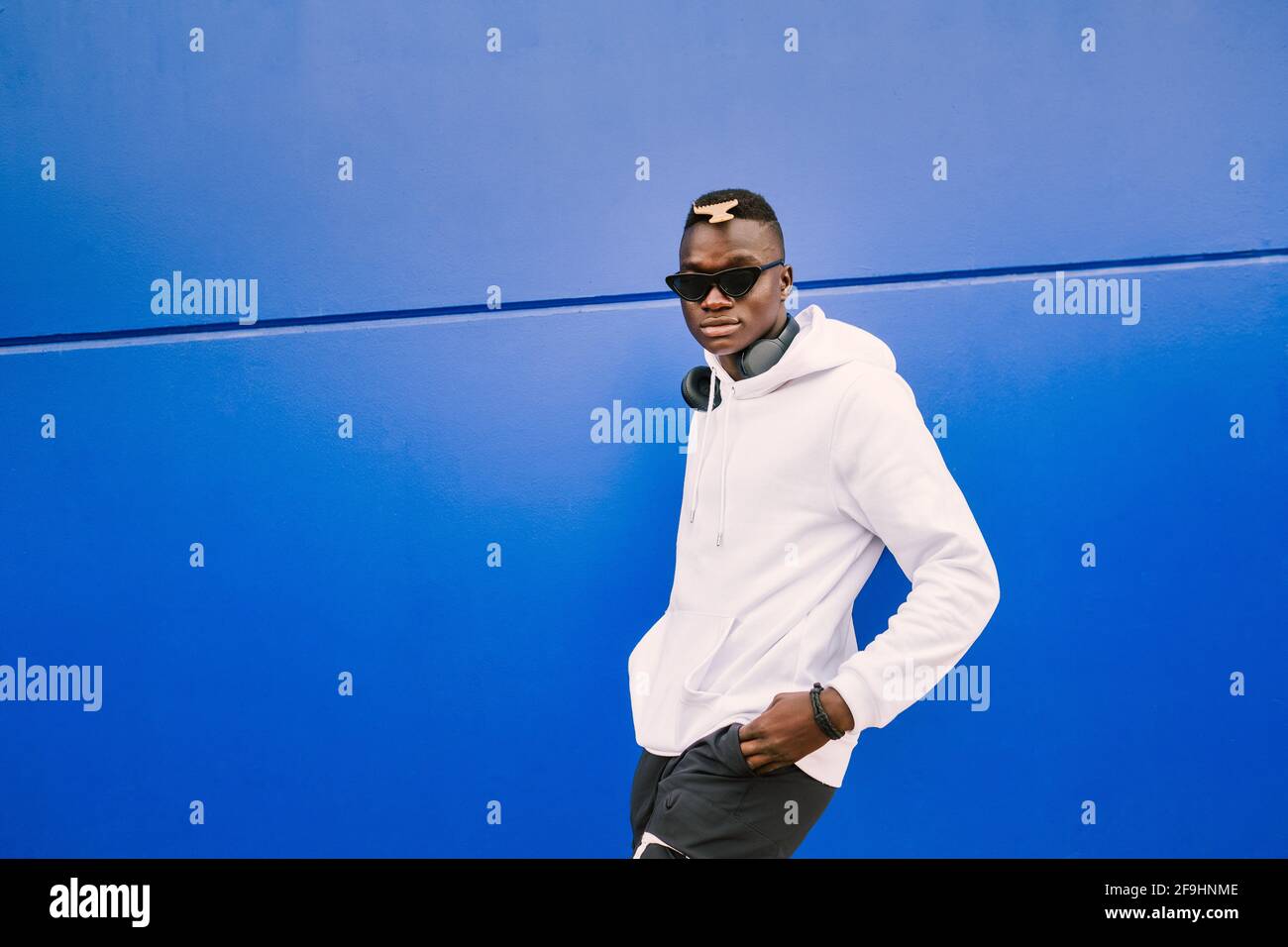 Young african black man against a blue wall looking at camera wearing a white sweatshirt and sunglasses with style and confidence Stock Photo
