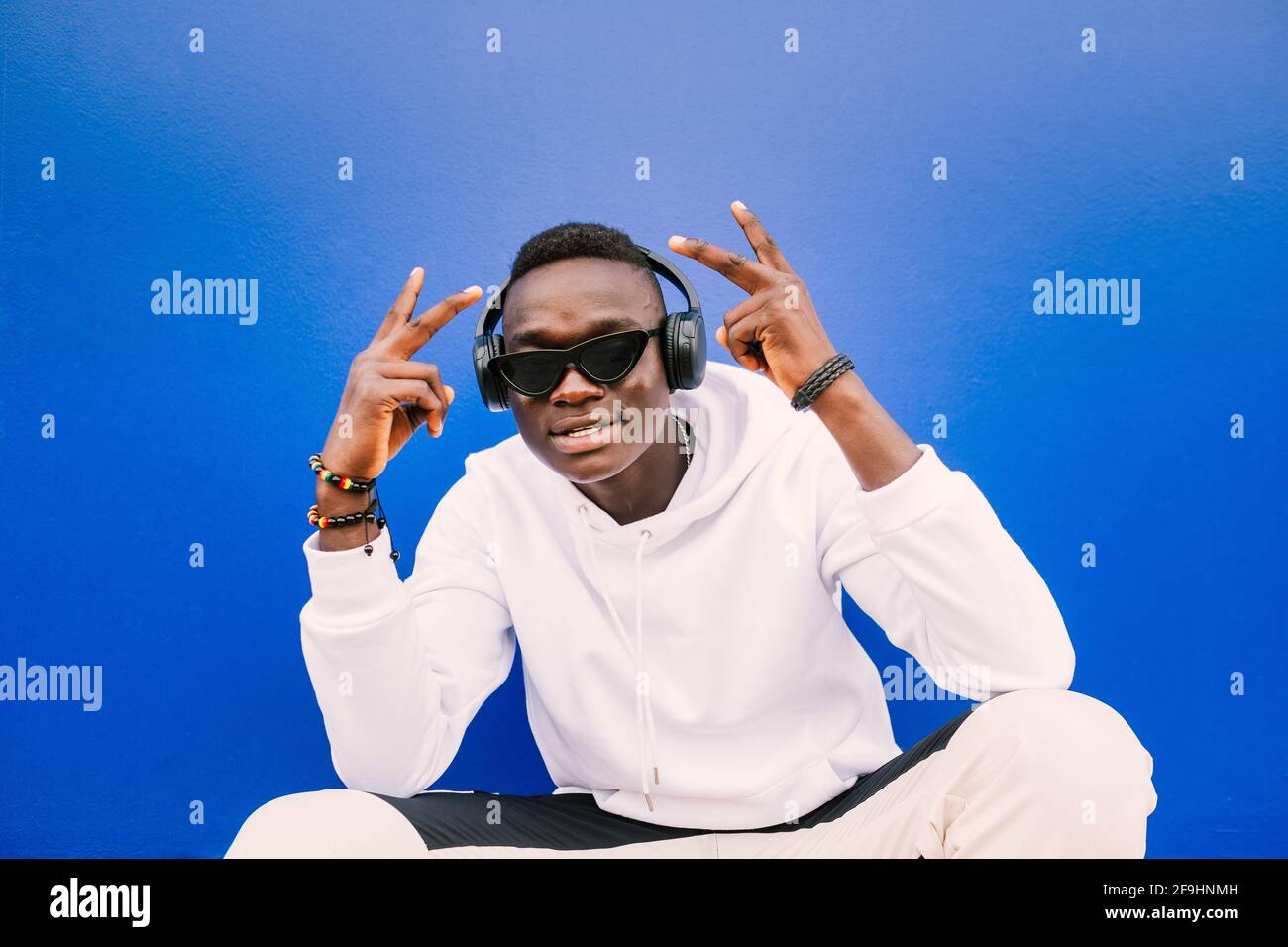 Young african black man seated against a blue wall looking at camera wearing a white sweatshirt and sunglasses doing the victory sign Stock Photo
