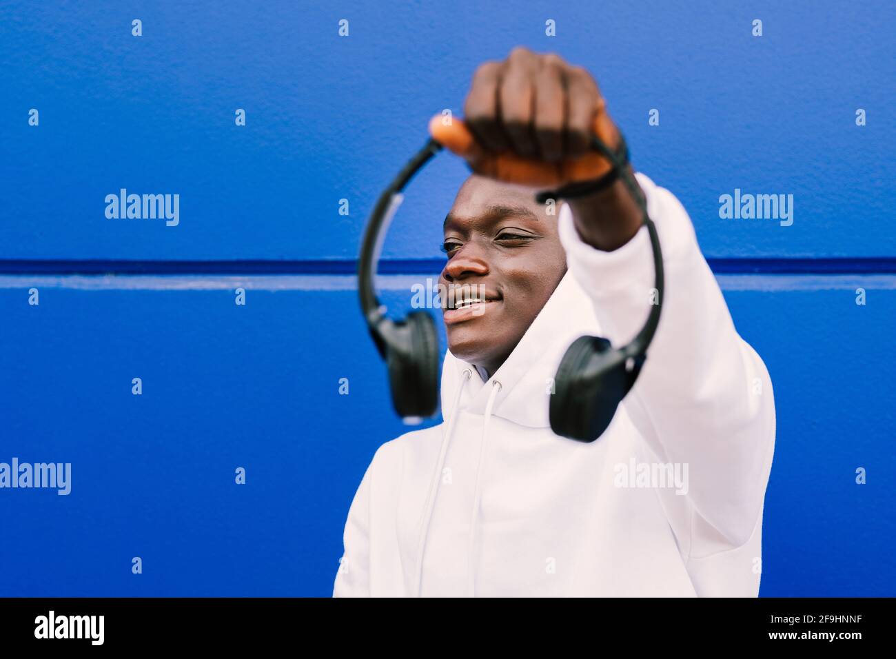 Young african black man against a blue wall wearing a white sweatshirt holding wireless headphones. Selective focus. Stock Photo