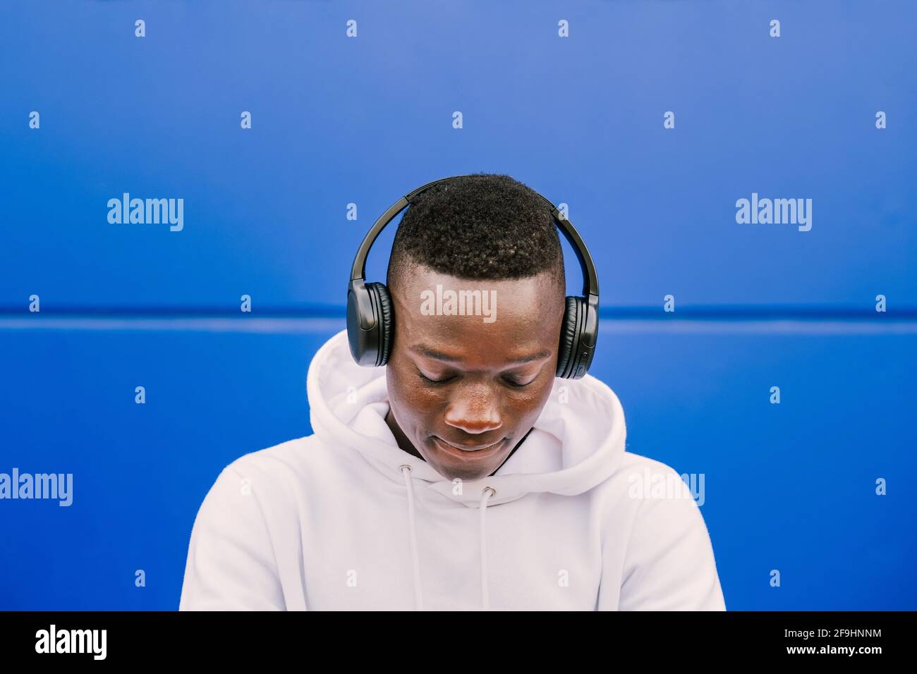Young african black man against a blue wall wearing a white sweatshirt listening music on wireless headphones while looking down Stock Photo