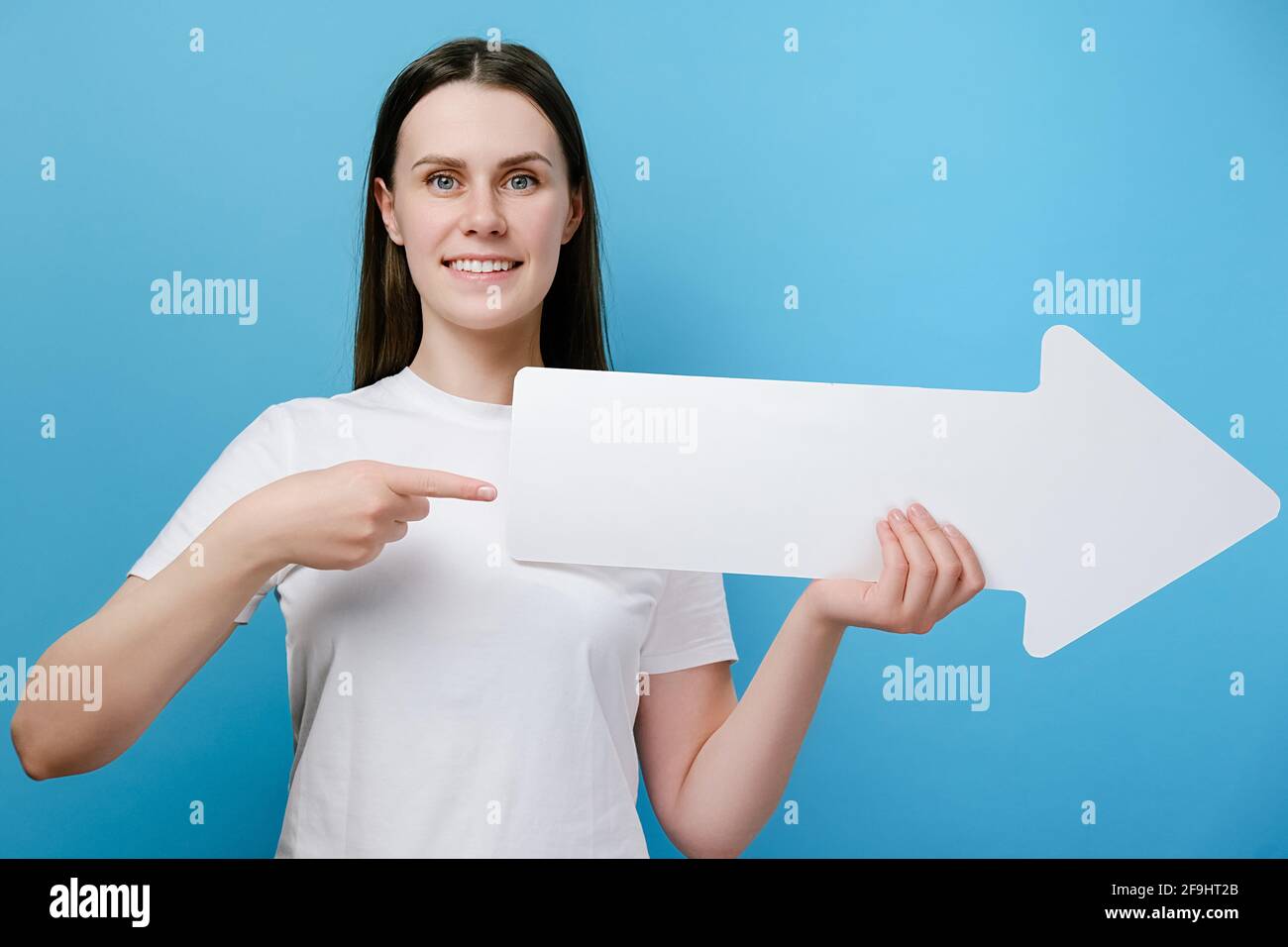 Excited caucasian young woman 20s smiling points fore finger on paper arrow aside, isolated over blue background with copy space for advertisement Stock Photo