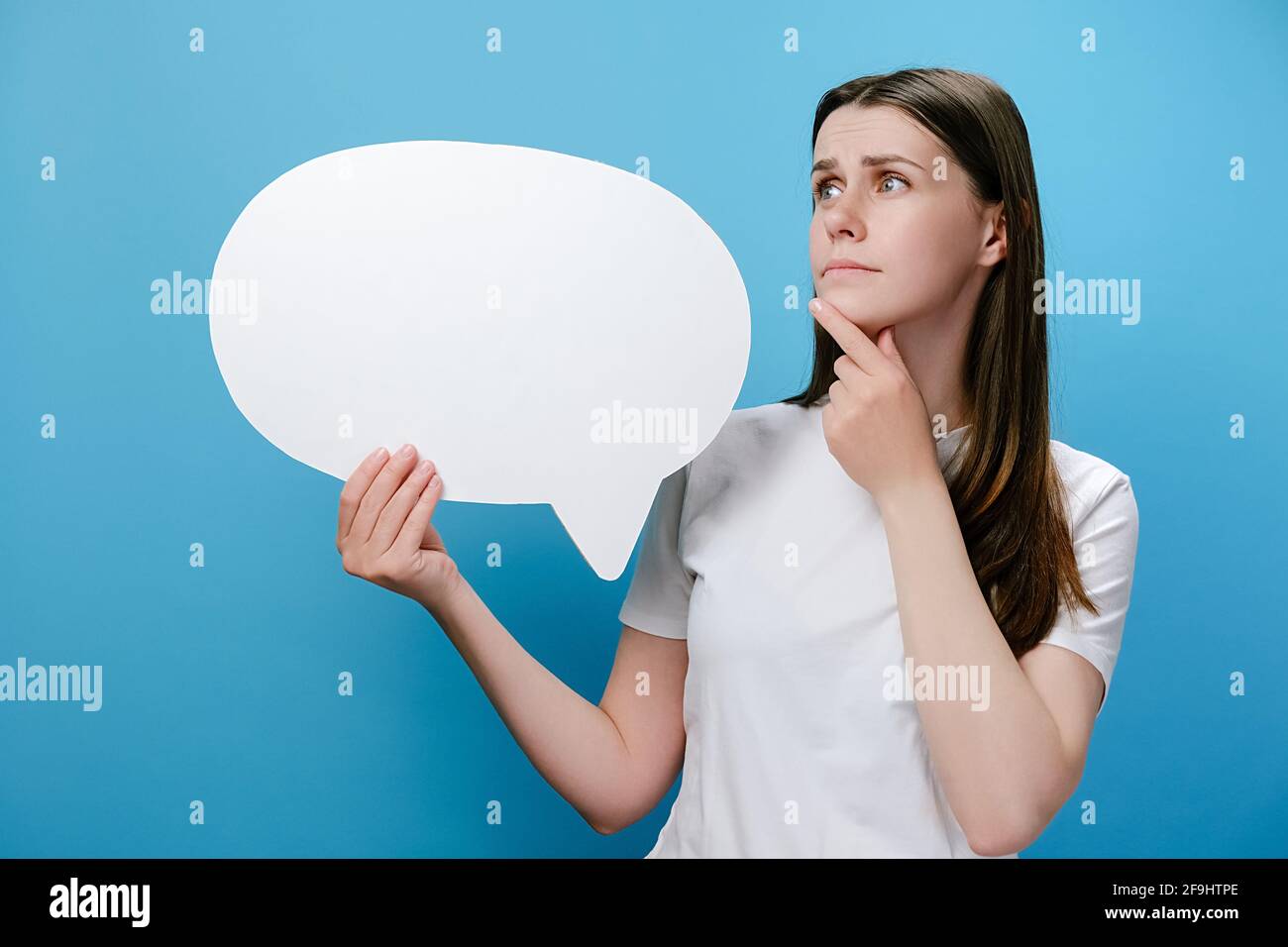 Worried pensive confused young female holding empty speech bubble, spreading arms put hand prop up on chin, dressed in white t-shirt Stock Photo