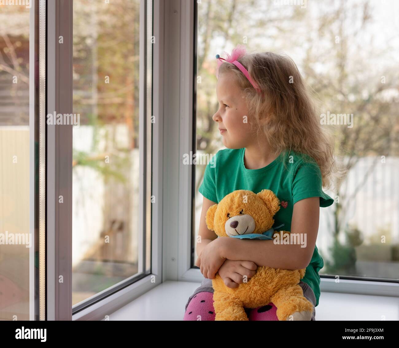 Cute little girl is sitting with her teddy bear near window and looking outside Stock Photo