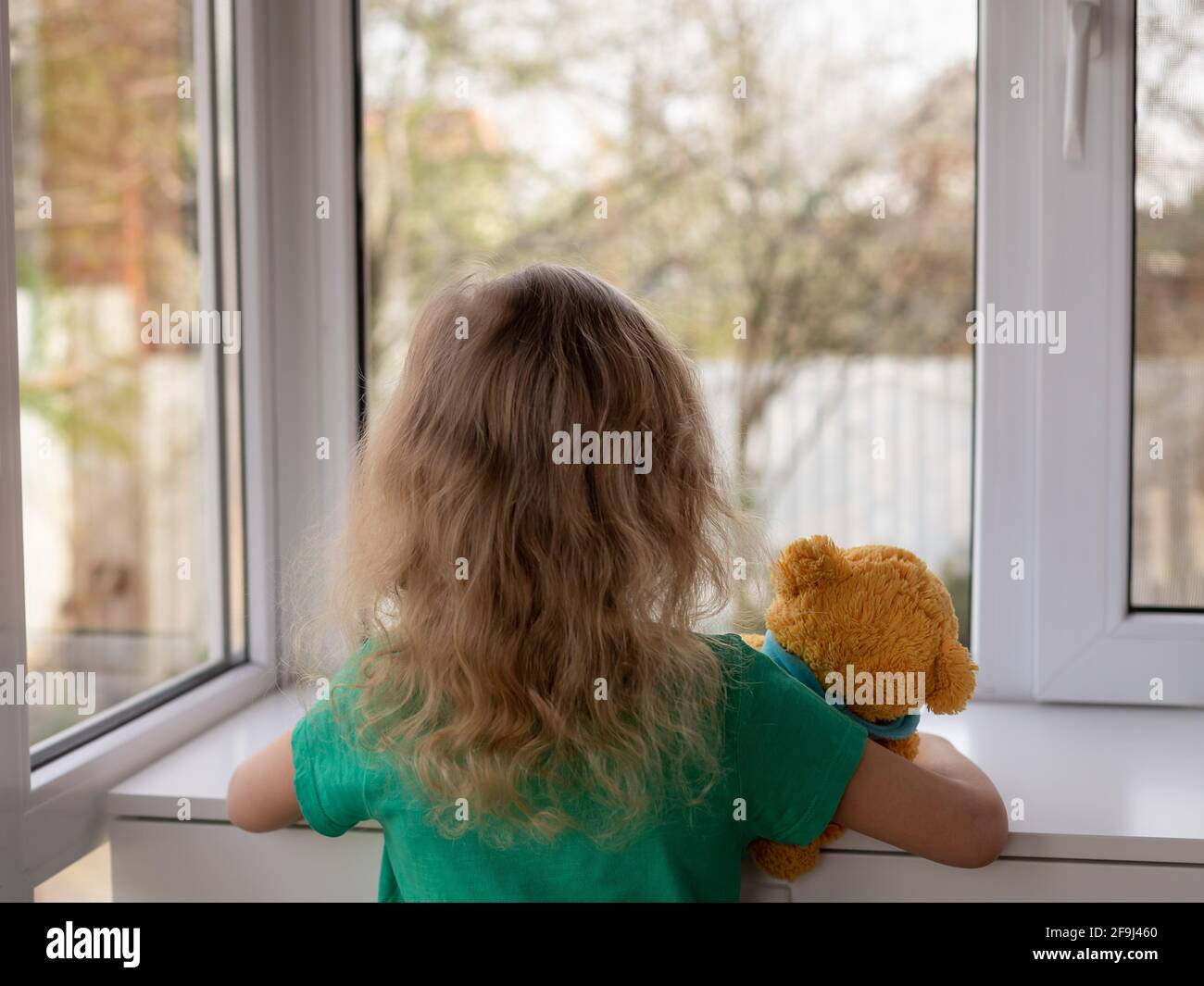 a little girl with her teddy bear looks out the window at the garden. stay home concept. selective focus Stock Photo