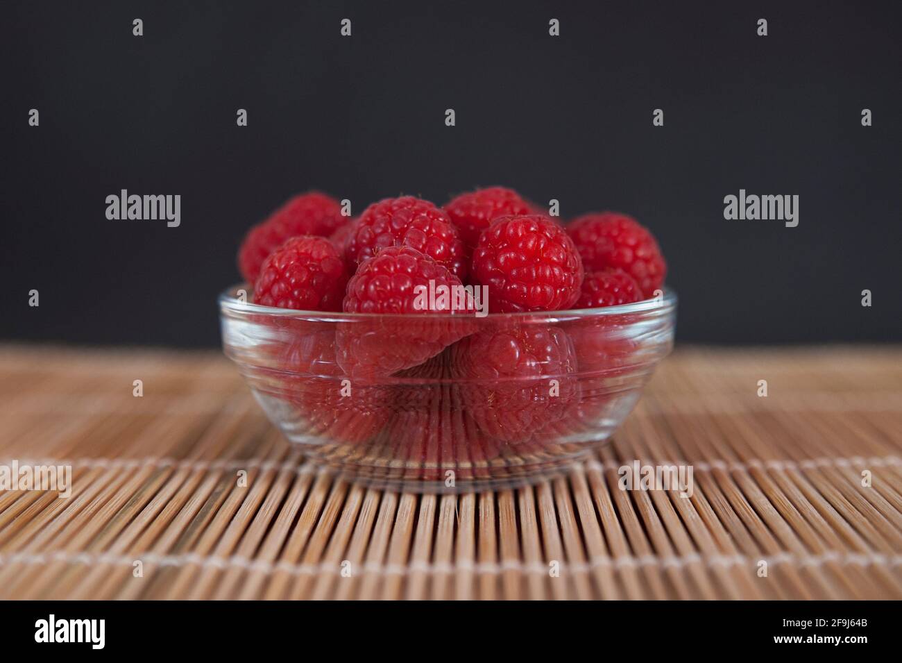 Centered, freshly picked raspberries in a bowl positioned on a bamboo mat against dark background, juicy, healthy and juicy fruit of genus Rubus Stock Photo