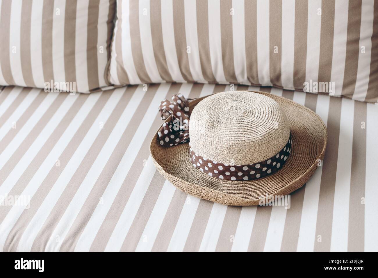 Straw hat with dotted silk ribbon laying on white and beige striped upholstered sofa. Classic furniture. Summer fashion accesories. Relaxation concept Stock Photo