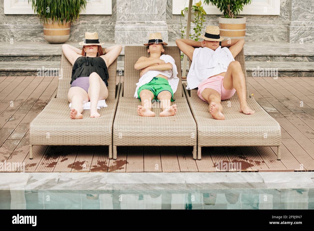 Father, mother and son covering faces with hat when sunbathing on chaise-lounges by swimming pool Stock Photo