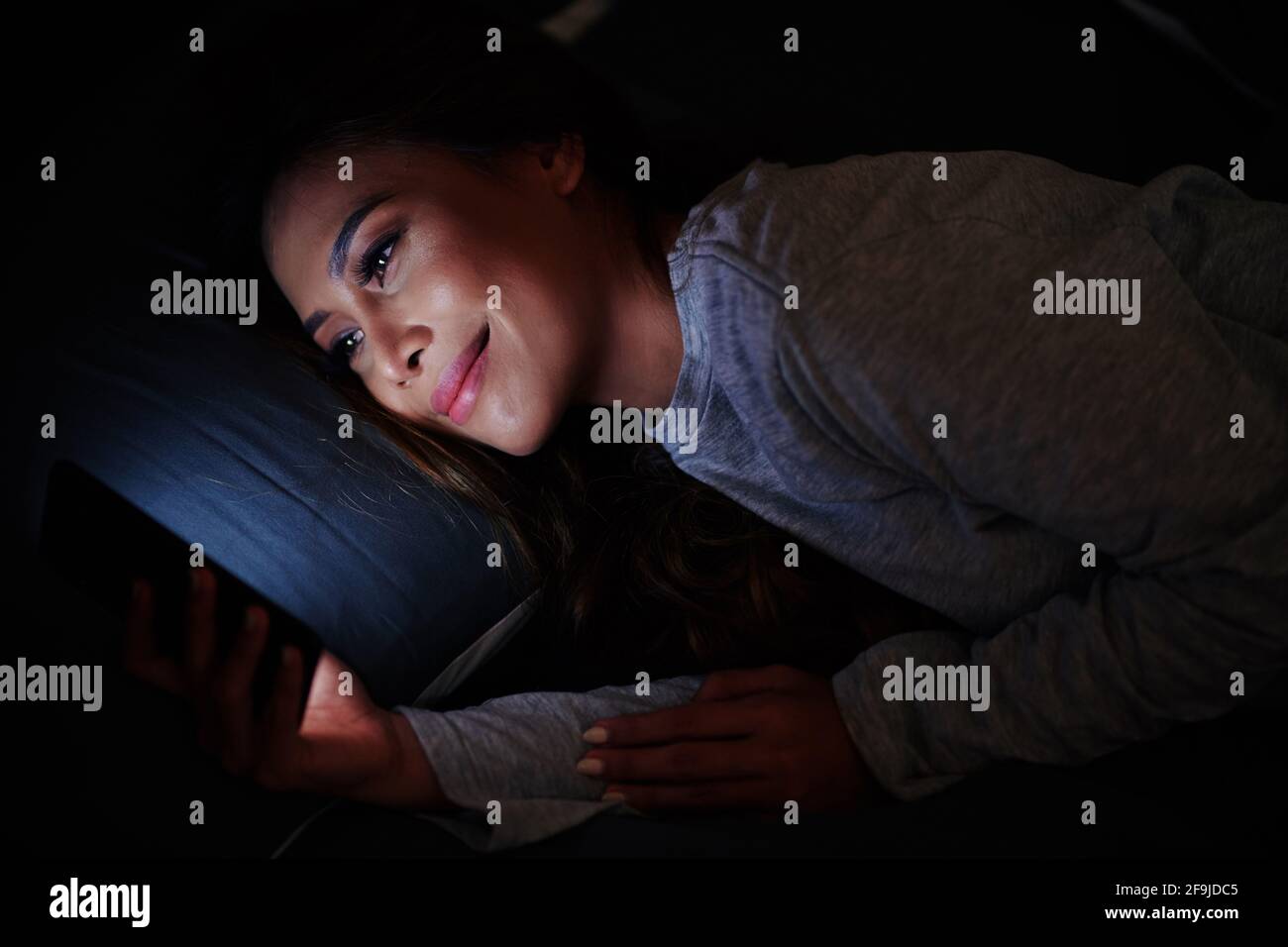 Smiling attractive young Asian woman lying on bed in dark room and watchong movie on glowing screen of smartphone Stock Photo