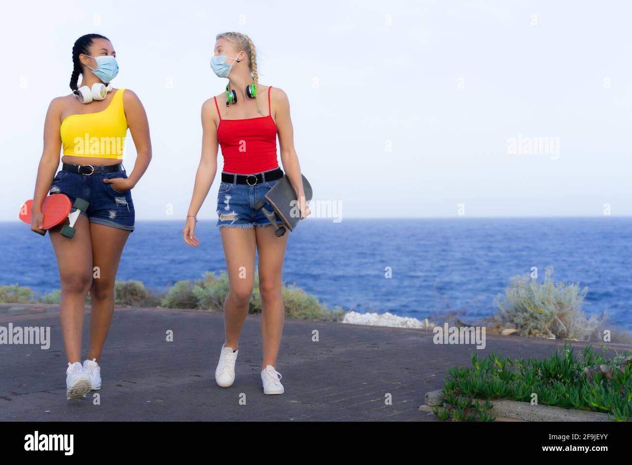 Young women walking, carrying a skateboards and looking at each other with protective mask for coronavirus. Friendship and covid concept. Stock Photo