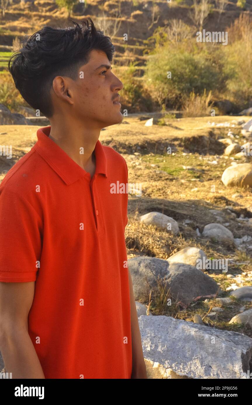 Portrait of a south asian young guy standing in outdoor and looking sideways, Young man with red color tshirt, A young adult guy with imperfect skin Stock Photo
