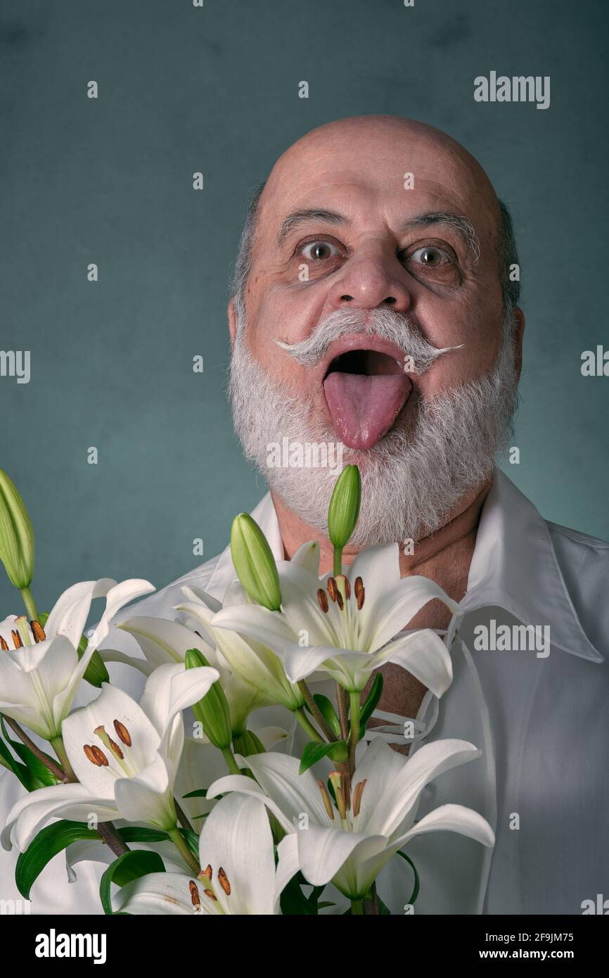 portrait of an elderly man, bald, with a gray beard and Latin appearance, wearing a white shirt and holding a bouquet of white lilies Stock Photo