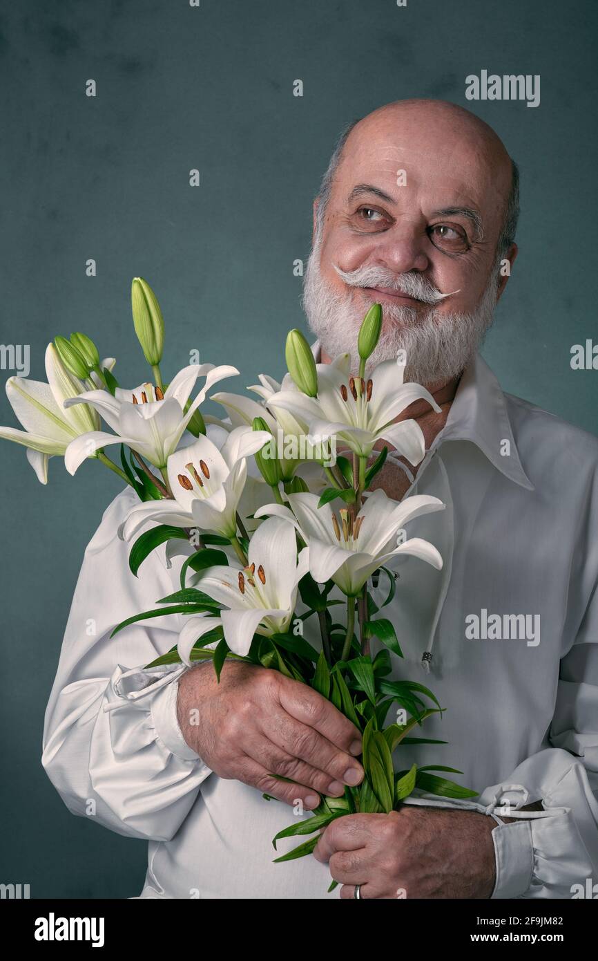 portrait of an elderly man, bald, with a gray beard and Latin appearance, wearing a white shirt and holding a bouquet of white lilies Stock Photo
