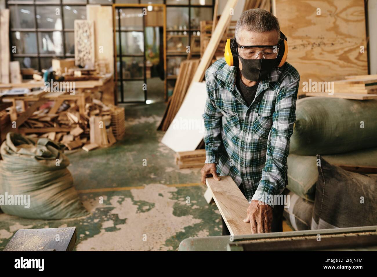 Asian carpenter in protective mask and earmuffs putting wooden board in thickness planer Stock Photo