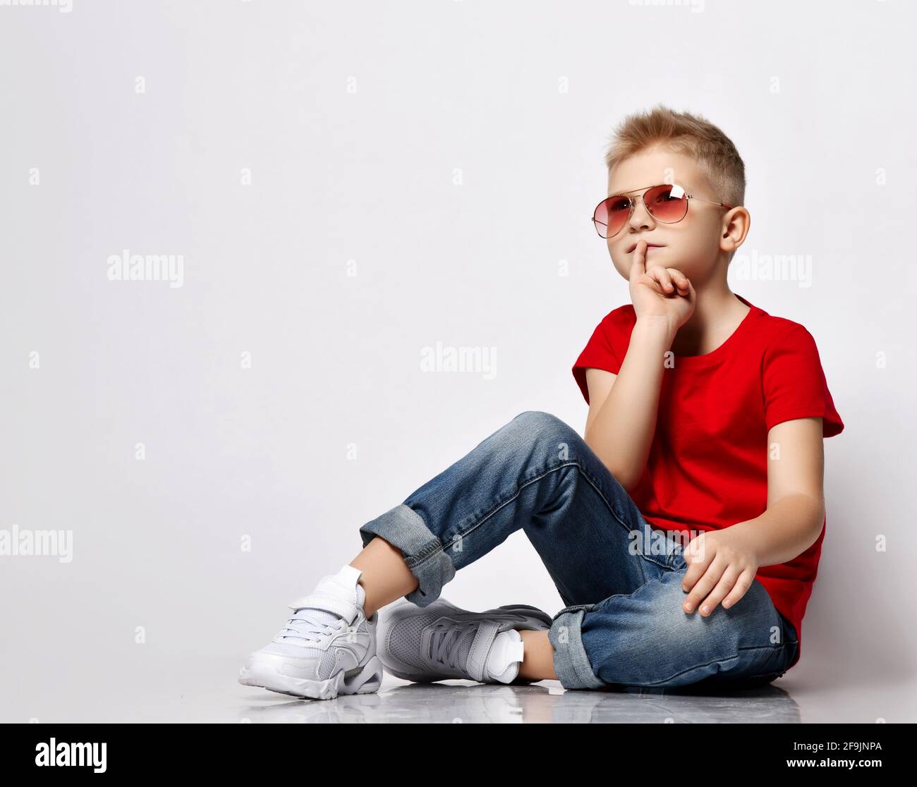 Stylish blonde kid boy leader in red t-shirt, blue jeans, white sneakers and sunglasses sits on floor thoughtful Stock Photo
