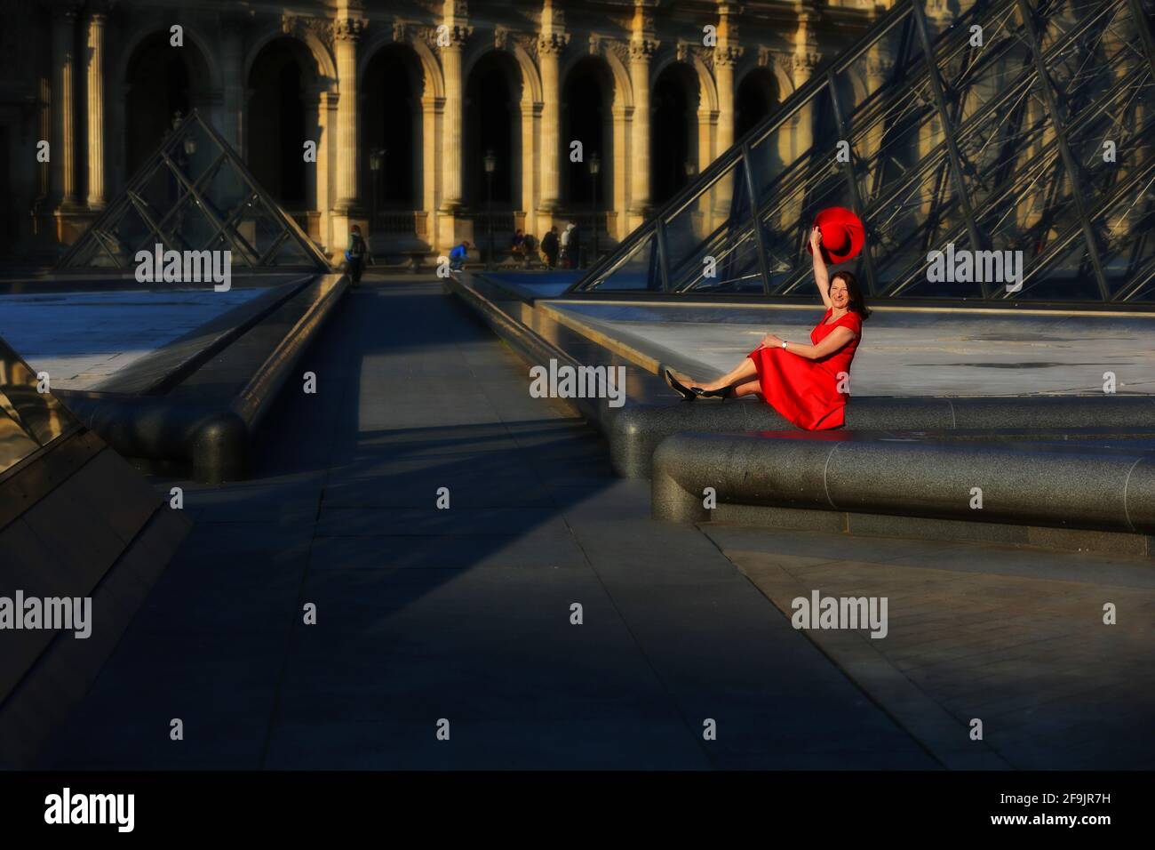 Paris, Frankreich, Louvre, sonniger Innenhof und Glaspyramide des Louvre in Paris in Frankreich mit sitzender Frau mit rotem Kleid und rotem Hut Stock Photo