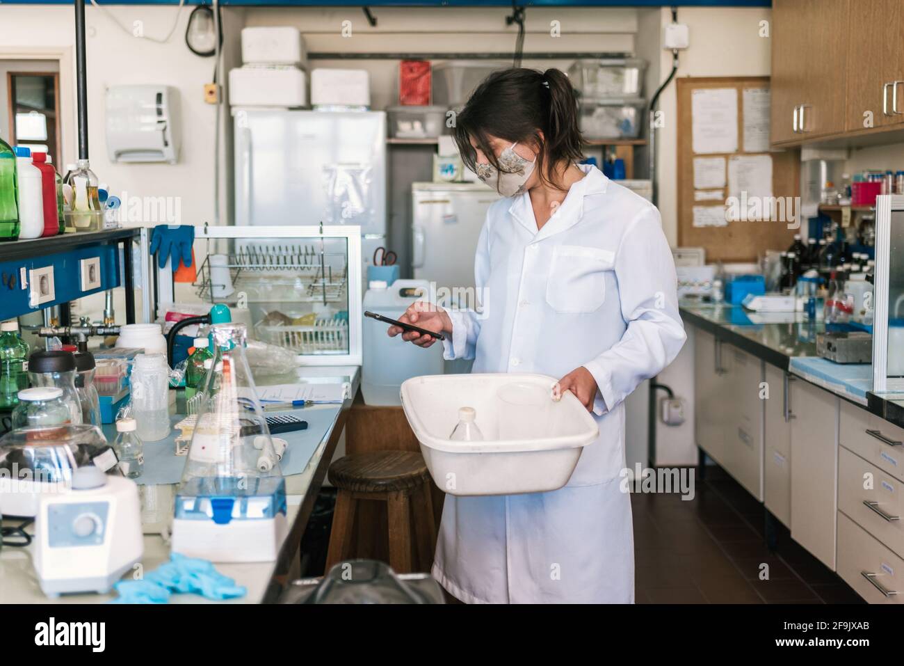 Side view of female scientist wearing face mask working in the laboratory with her mobile phone. Stock Photo