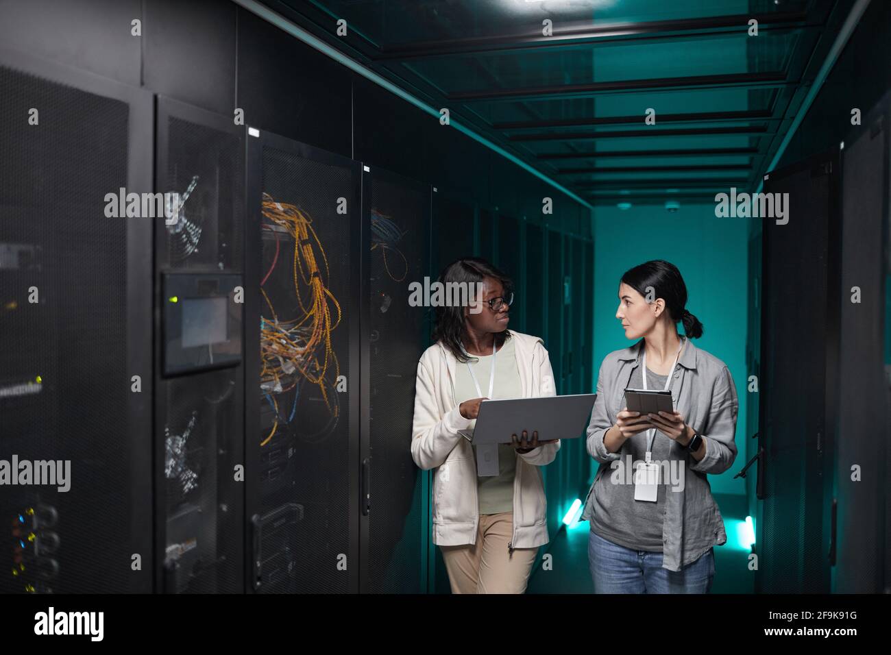 Portrait of two young women using laptop in server room while setting up supercomputer network, copy space Stock Photo