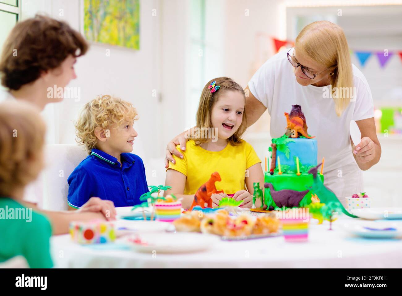 Kids birthday party. Dinosaur theme cake. Little girl blowing candles and opening gifts. Children event. Decoration for dino themed celebration. Stock Photo