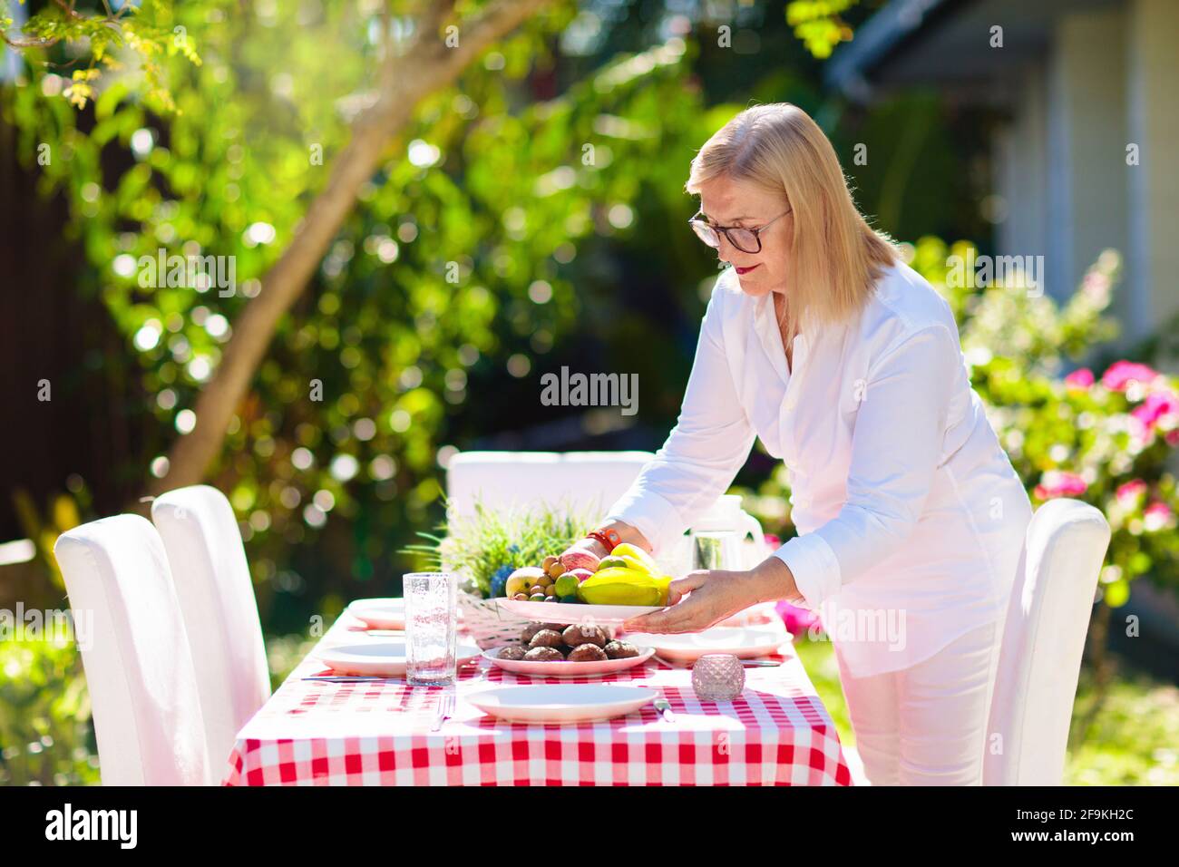 Woman setting table outdoors. Garden summer fun. BBQ in sunny backyard. Senior lady at lunch. Party decoration. Female with fruit outdoor. Stock Photo