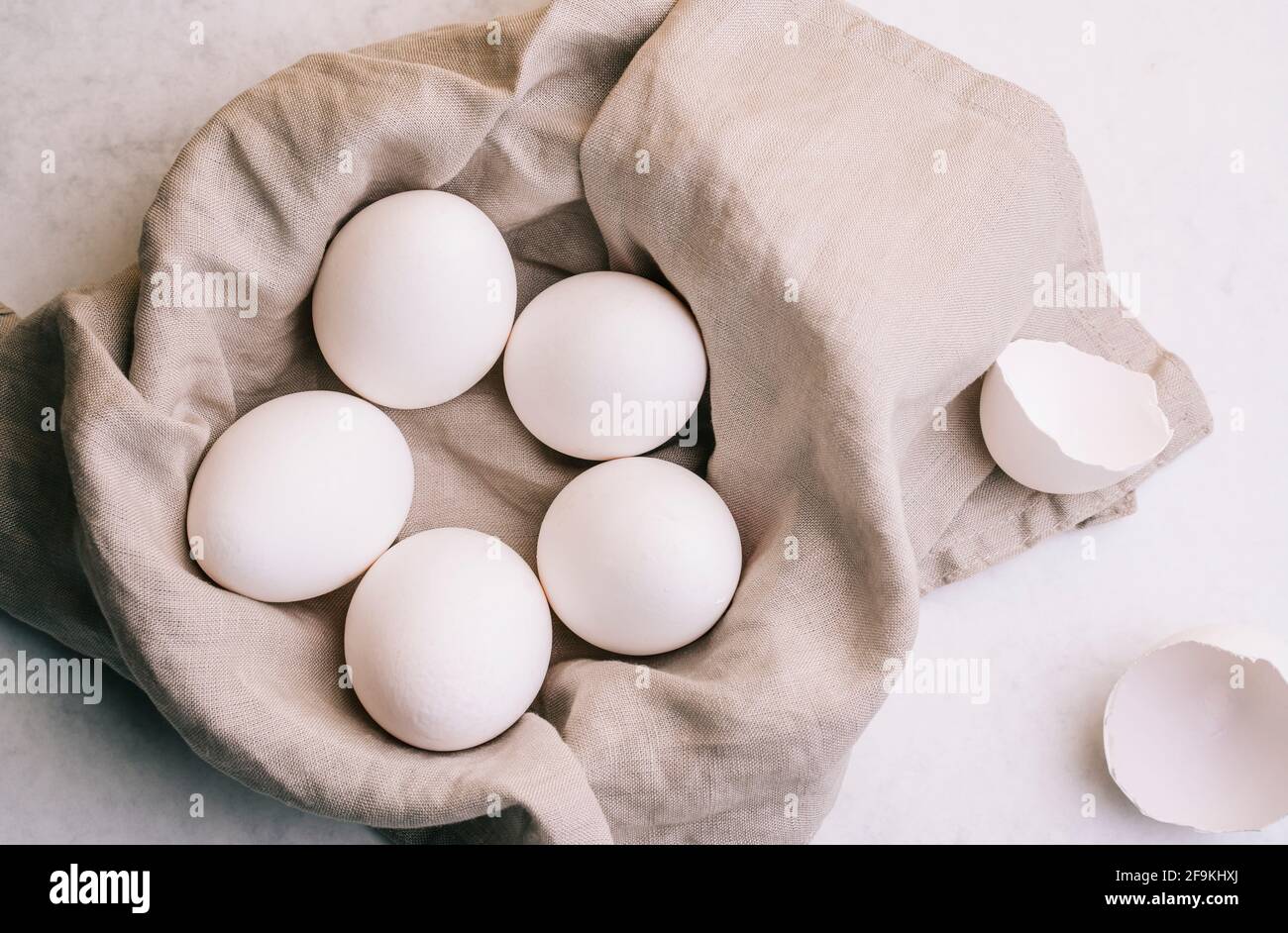 Basket of white eggs on a gray linen cloth with two shells beside it on white marble background illuminated by the light from the window Stock Photo