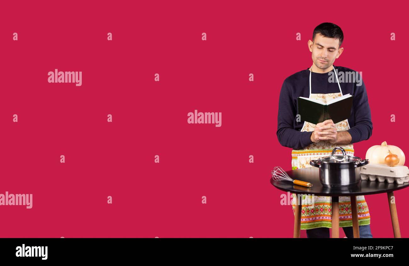 Serious handsome young man wearing professional baker apron reading cooking recipe book surprised, smiling and thinking on red background with empty Stock Photo