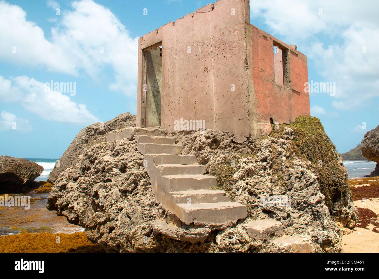 Ruins of a dirty pink concrete building on a rock formation on Bathsheba Beach, Barbados, during the day. Thick clouds, blue skies. Stock Photo