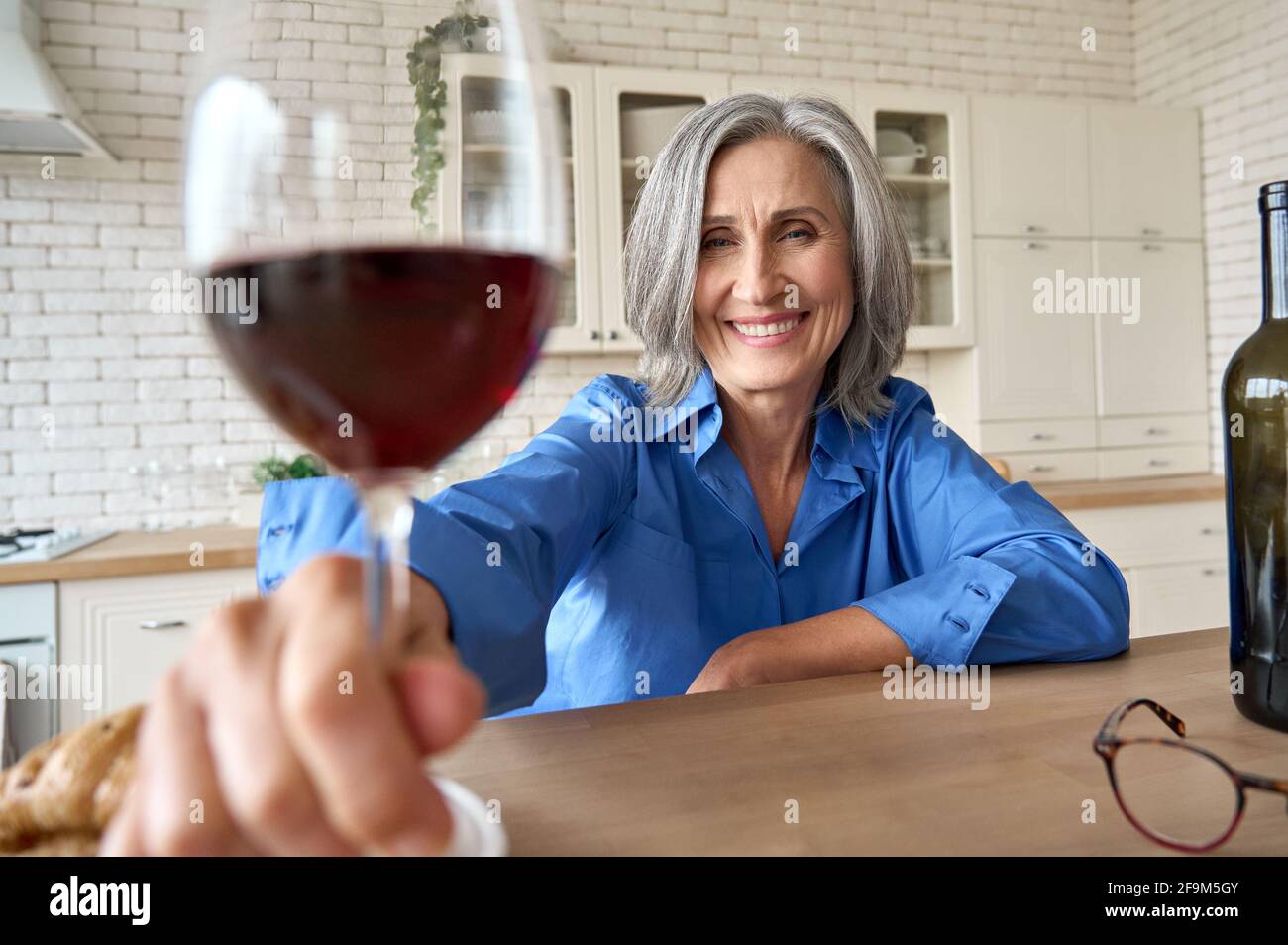Middle age 60s woman sitting at kitchen drinking wine online with family. Stock Photo