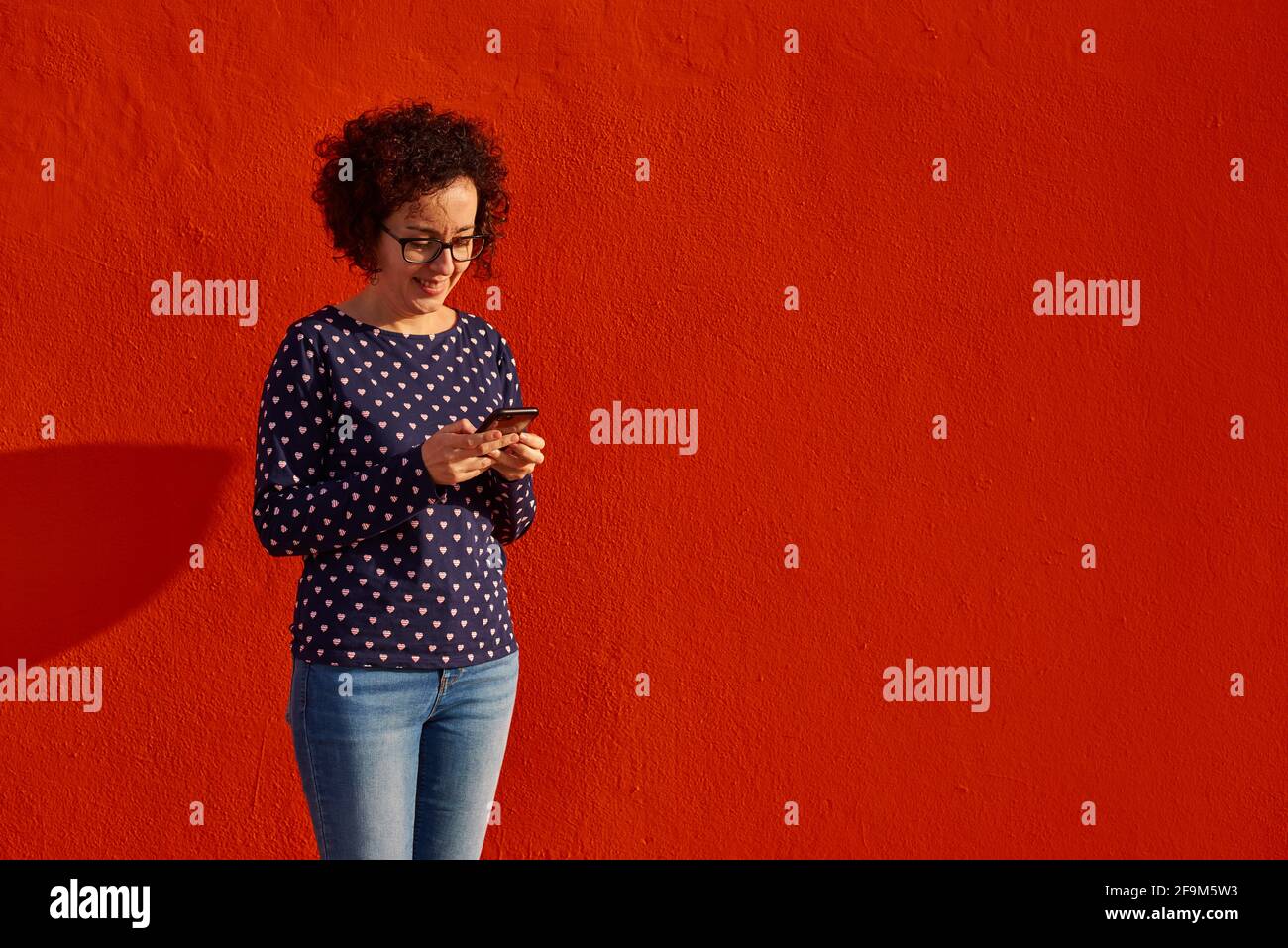 Front view of a happy woman standing against a bright red wall using her smartphone. High quality photo. Stock Photo