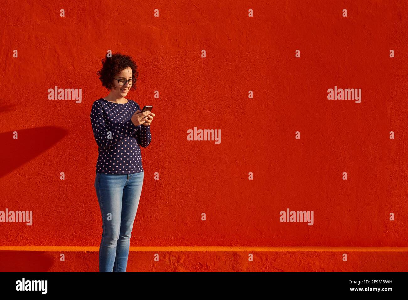 Front view of a happy woman standing against a bright red wall using her smartphone. High quality photo. Stock Photo