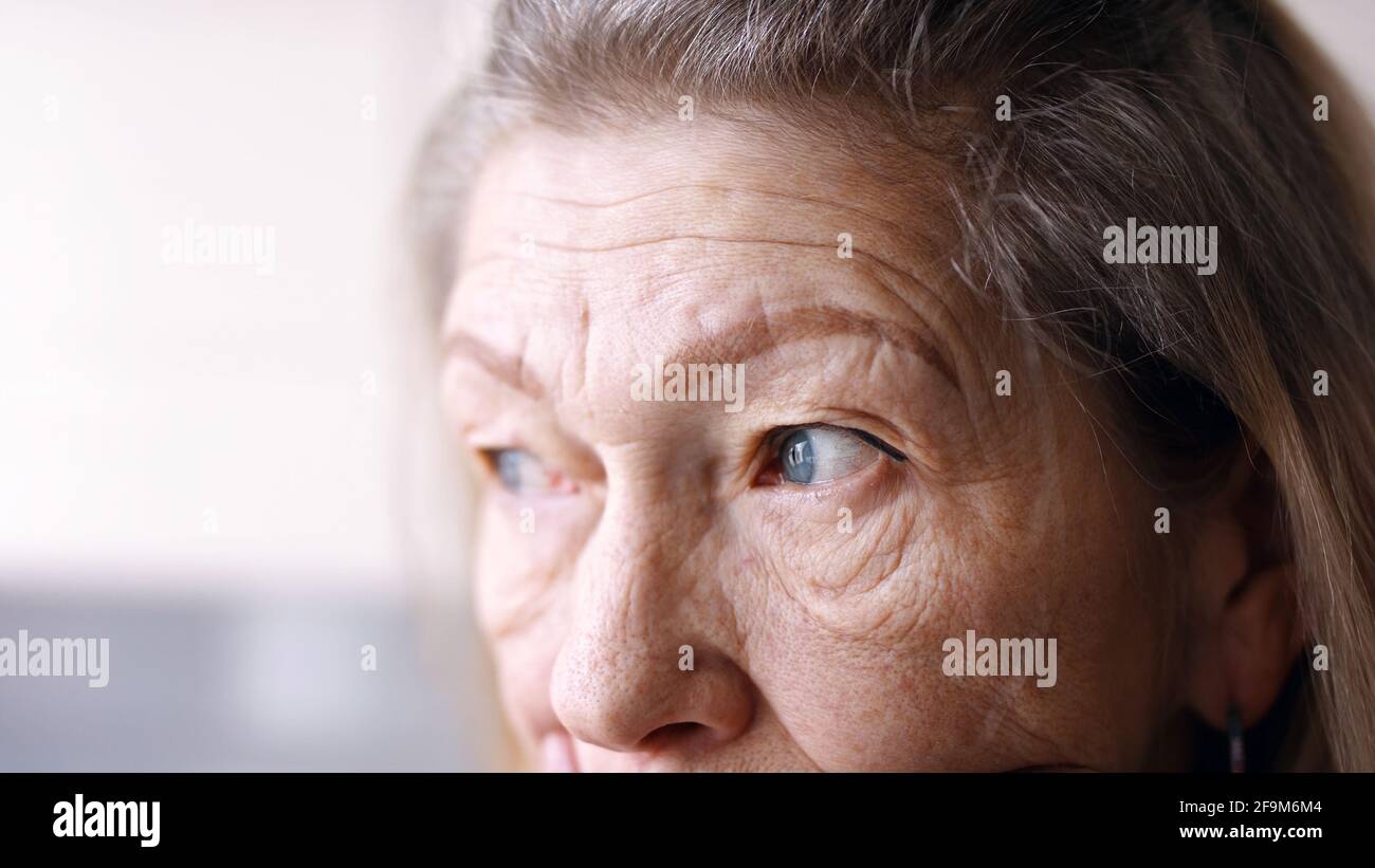 Close up on blue sad eyes of lonely elderly woman looking through the window. High quality photo Stock Photo