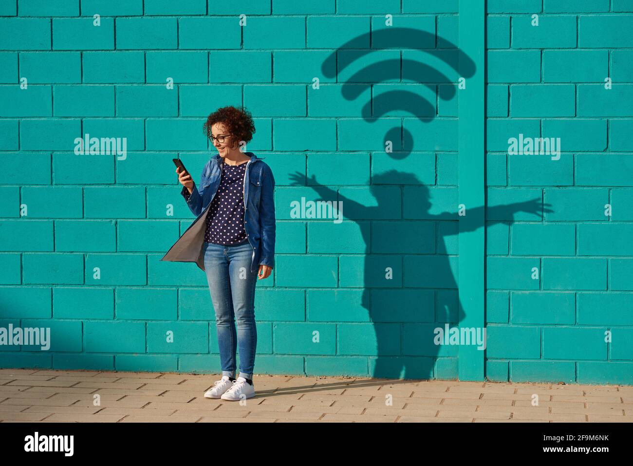 Front view of a happy woman standing against a bright blue wall using her smartphone with a shadow drawing a wifi signal Stock Photo