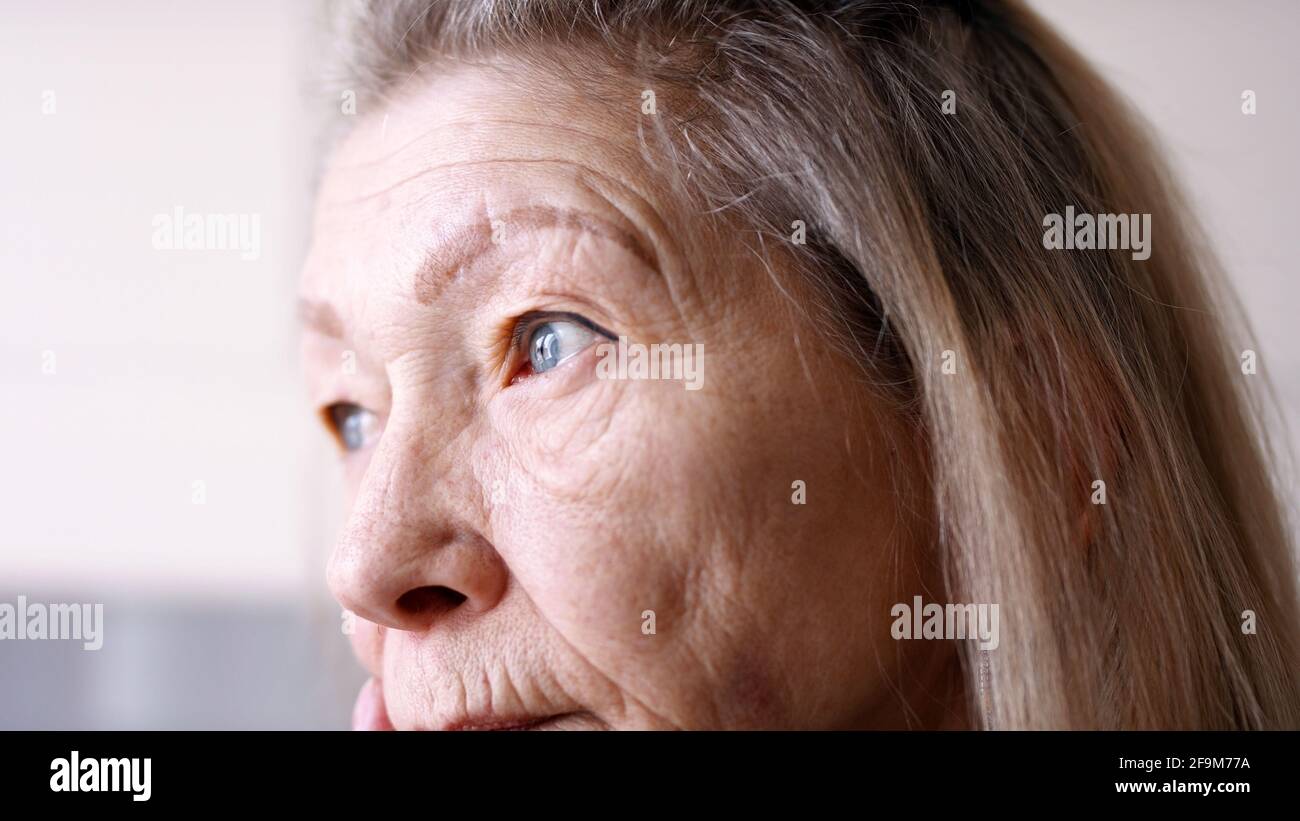 Close up on blue sad eyes of lonely elderly woman looking through the window. High quality photo Stock Photo