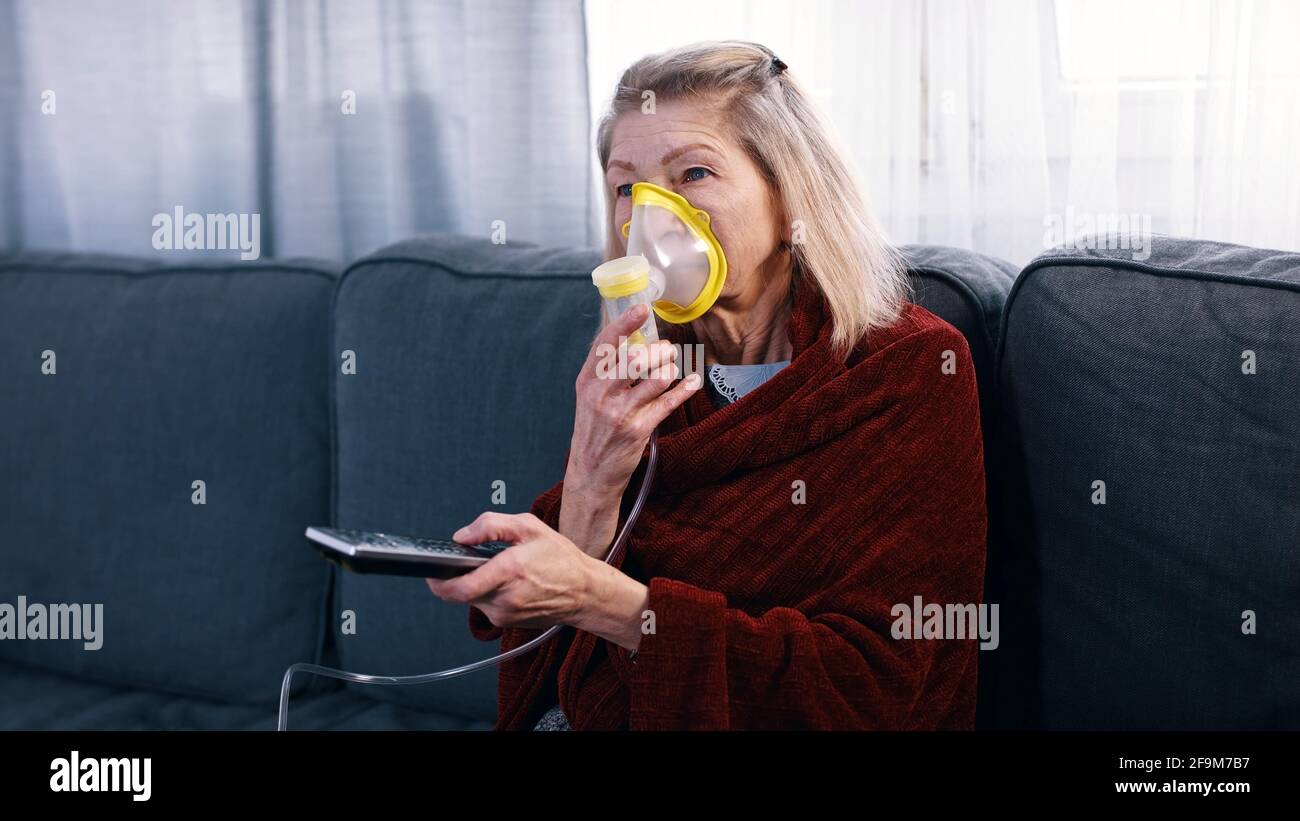 Elderly woman using oxygen mask while changing channels on tv at home. High quality photo Stock Photo