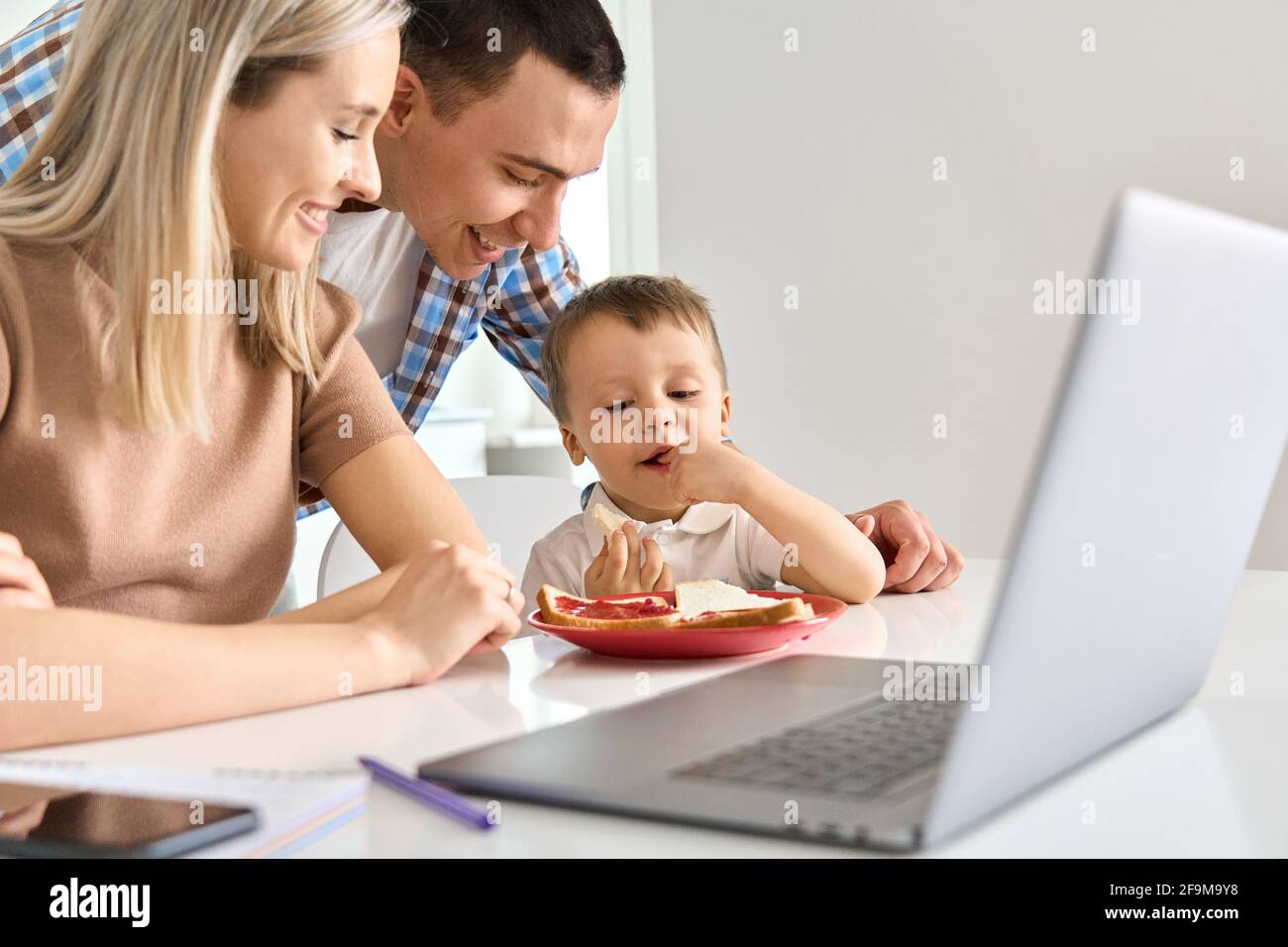 Happy young family with cute kid son eating toasts using laptop in kitchen. Stock Photo