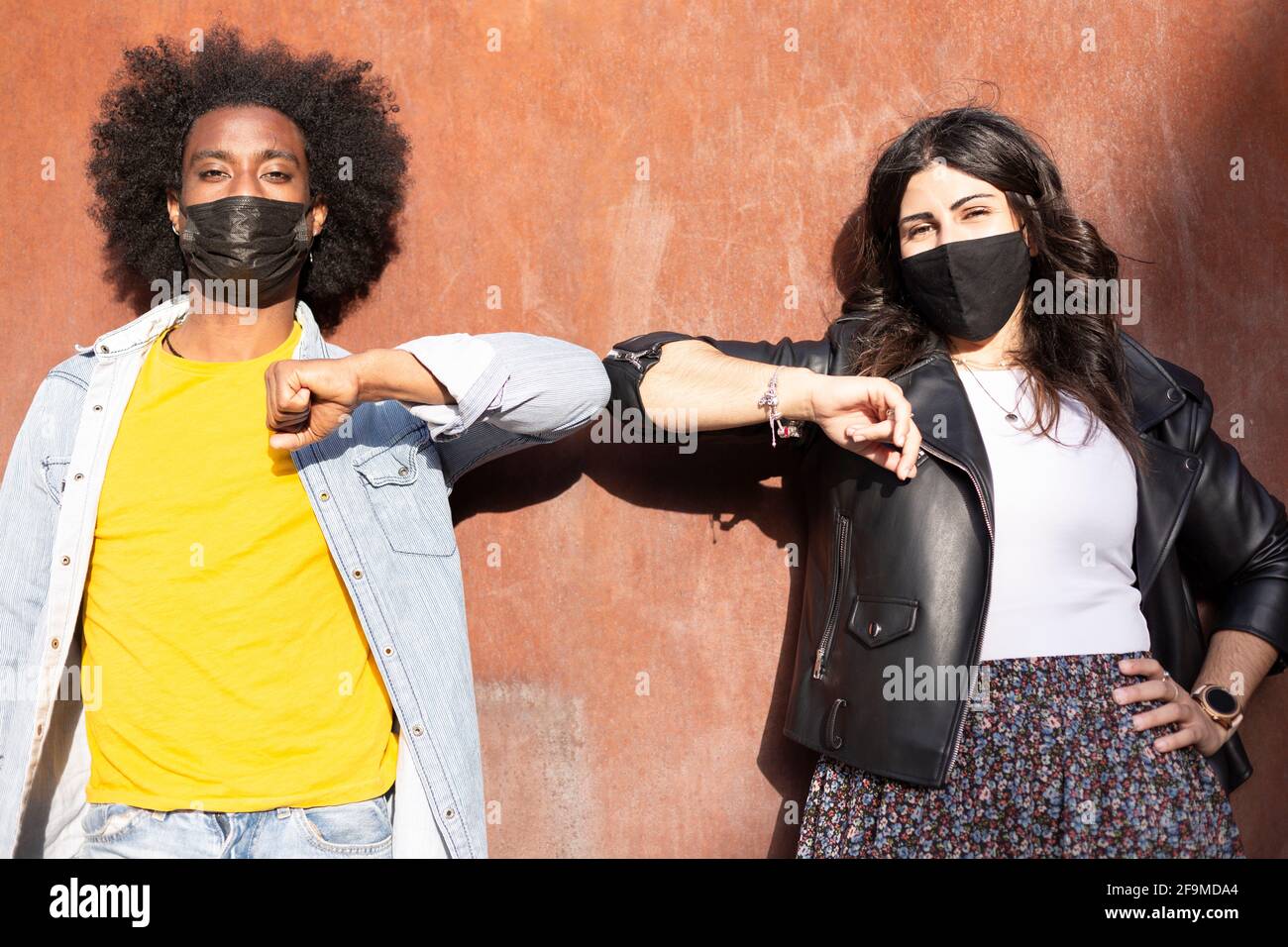 Couple of multiethnic friends wearing protective masks greet each other by bumping elbows and keeping the recommended distance to avoid Covid-19 virus Stock Photo
