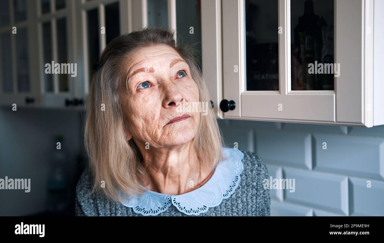 Sad lonely old gray haired woman looking through the window. Vulnerable person in quarantine. High quality photo Stock Photo