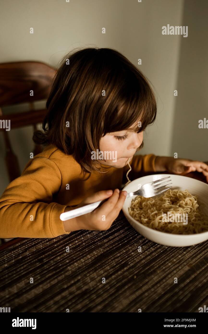 Young girl eating noodles in a yellow sweater at her kitchen table Stock Photo