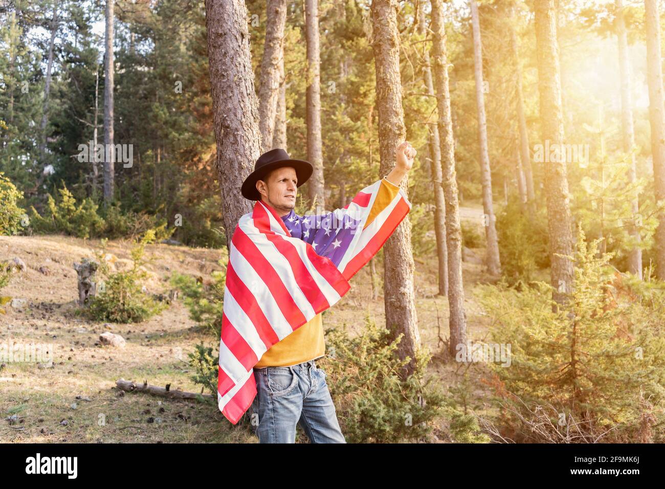 Young patriotic man in cowboy hat holding United States American flag and raising fist and arm in forest.Beautiful woodland landscape at sunset.Indepe Stock Photo