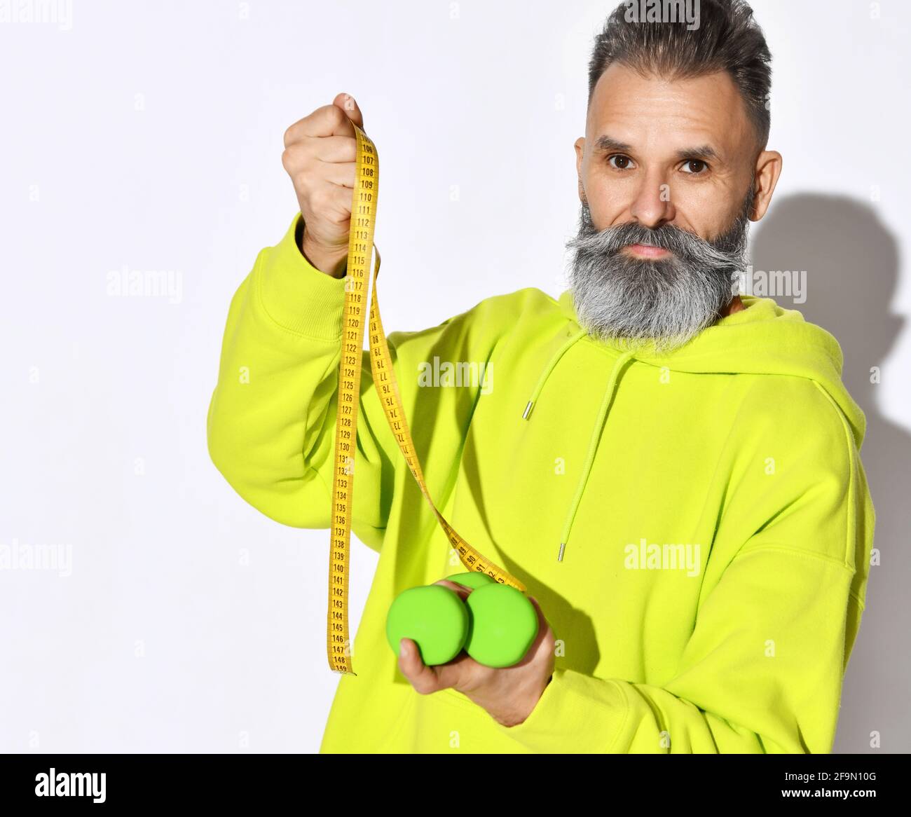 Mature hipster old brutal bearded man in yellow hoodie hold green dumbbells and tape measure with white stylish beard on white Stock Photo