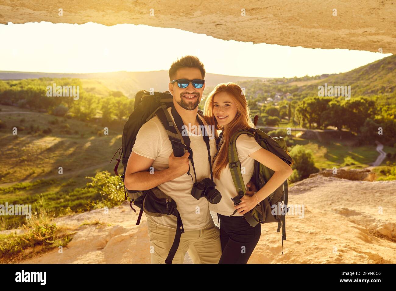 Happy young couple of hikers standing in mountains cave at sunset. Tourists mountaineering on summer vacation Stock Photo