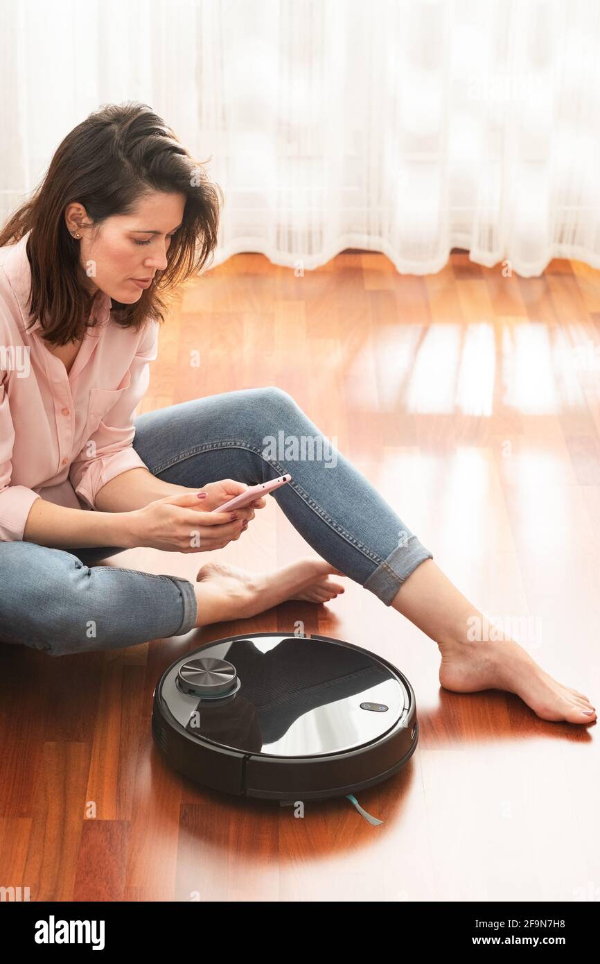 Young caucasian woman using automatic vacuum cleaner to clean the floor, controlling smart machine housework robot with smart phone at home Stock Photo