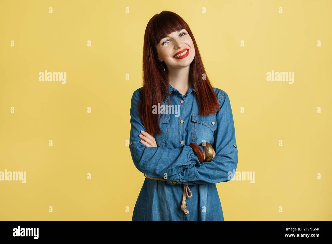 Pretty young red haired woman with bright red lipstick, wearing denim jeans  dress, posing with arms crossed on yellow background Stock Photo - Alamy